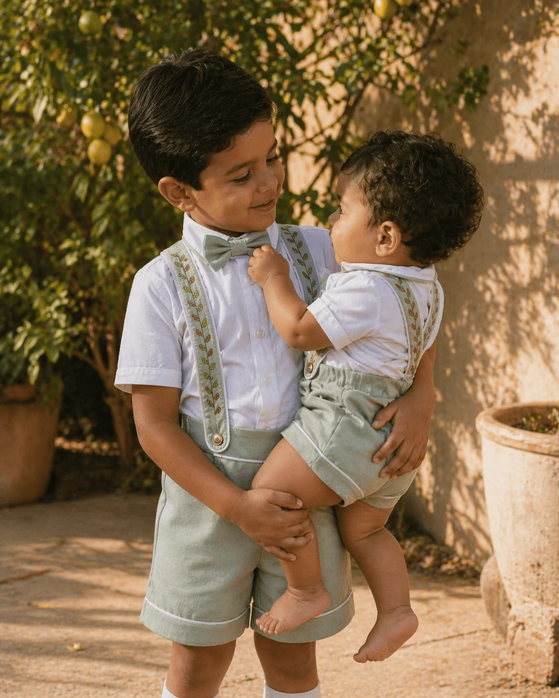 Irmão maior carregando o bebê no colo — ambos vestindo Conjunto Versátil verde sage — a mesma cliente que comprou o Body Camisa aos 6 meses volta pra encomendar a Camisa Social quando o filho sai da fralda
